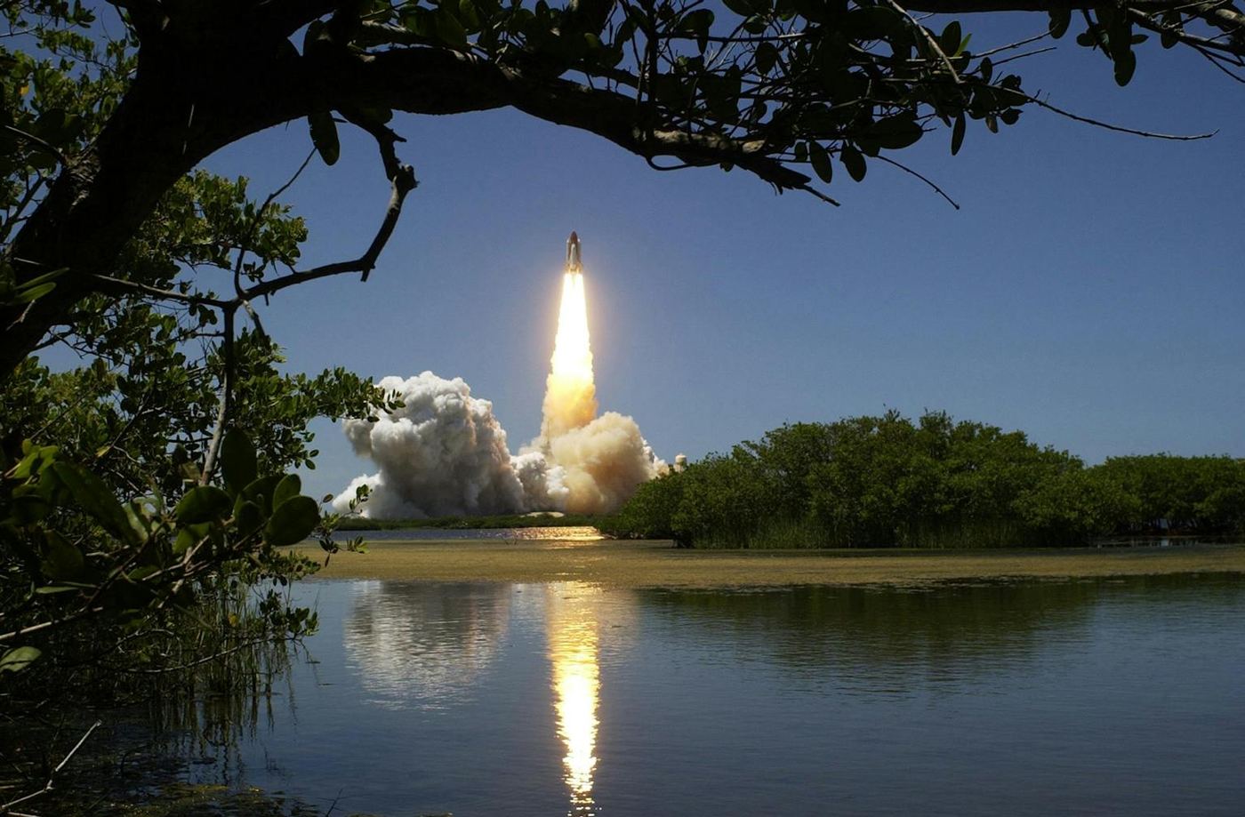 A dramatic rocket launch with smoke and flames reflected in calm waters and framed by trees