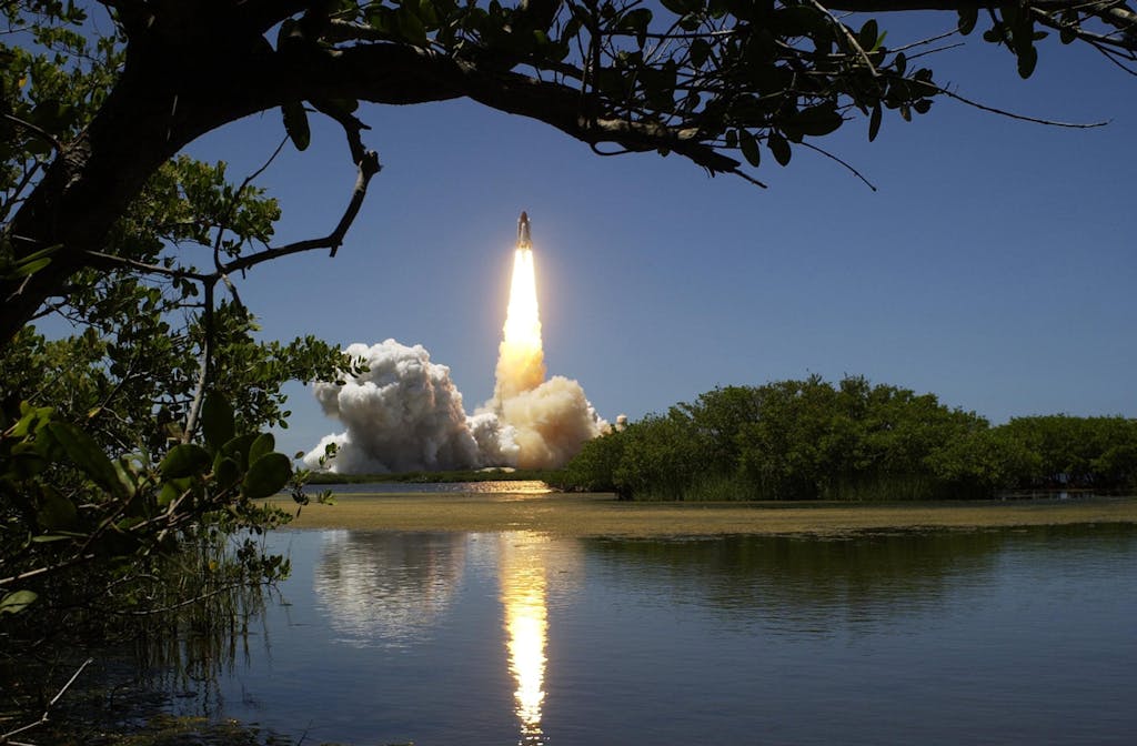 A dramatic rocket launch with smoke and flames, reflected in calm waters and framed by trees.